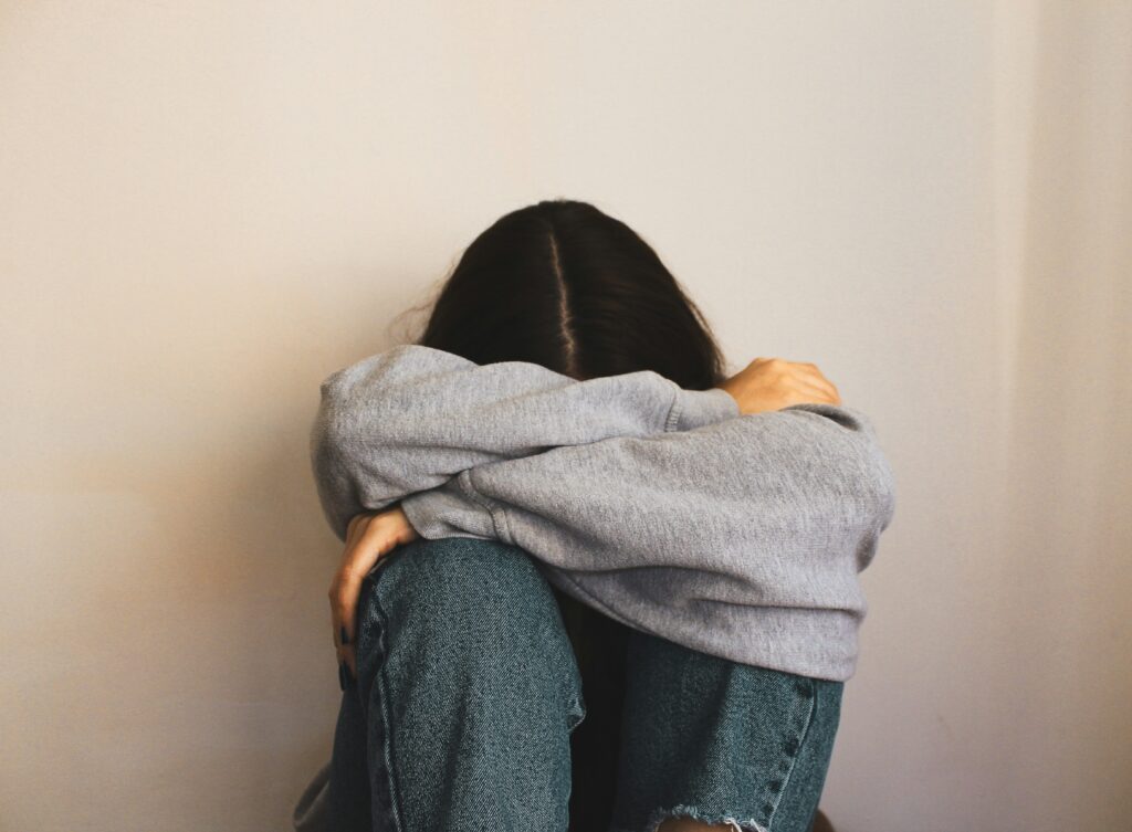 Person in grauem Pullover sitzt mit angezogenen Knien und verstecktem Gesicht an einer Wand, wirkt traurig oder überfordert.