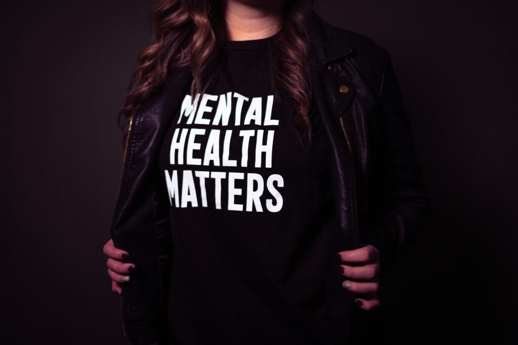 Person trägt ein schwarzes T-Shirt mit der weißen Aufschrift „Mental Health Matters“ und eine schwarze Lederjacke, der obere Teil des Gesichts ist nicht sichtbar.