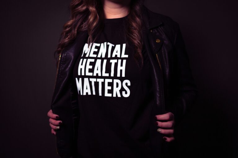 Person trägt ein schwarzes T-Shirt mit der weißen Aufschrift „Mental Health Matters“ und eine schwarze Lederjacke, der obere Teil des Gesichts ist nicht sichtbar.