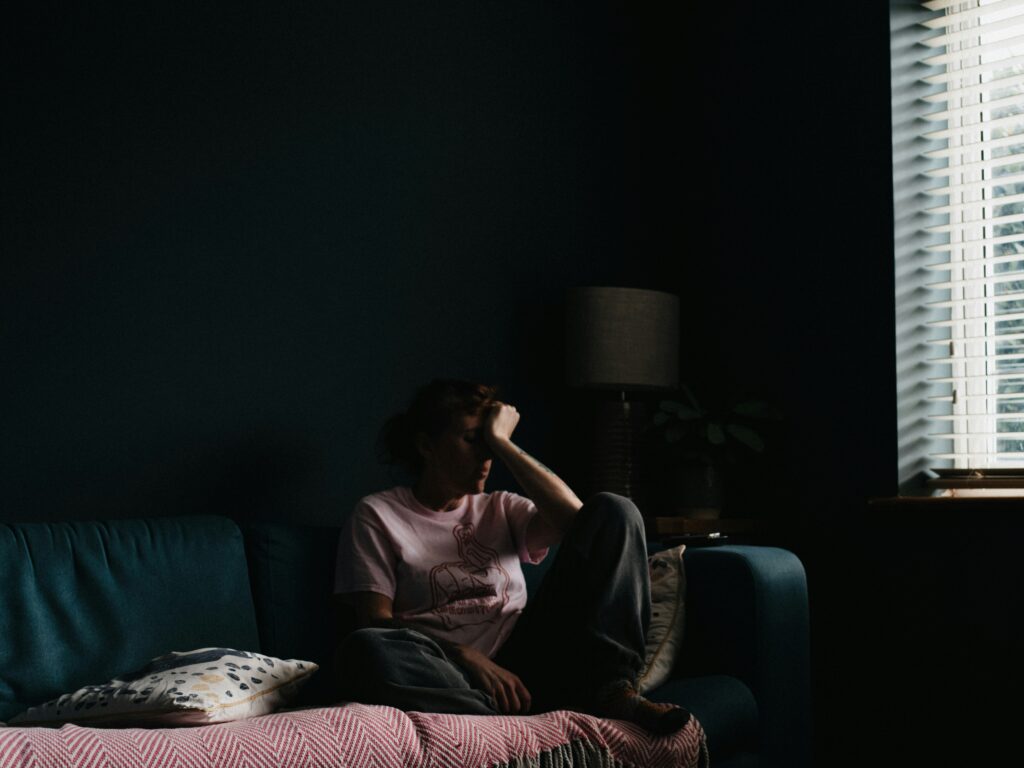 Die Frau sitzt allein und nachdenklich auf einem Sofa in einem dunklen Raum. Dies symbolisiert soziale Ängste und emotionale Überforderung im Studium.