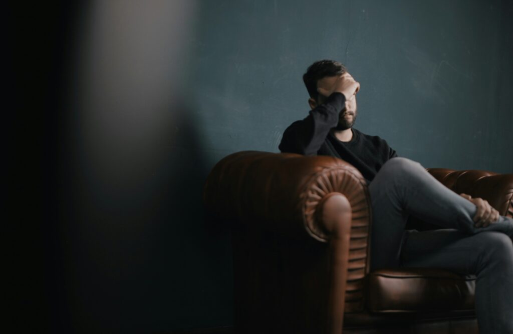 nik-shuliahin-BuNWp1bL0nc-unsplash Mann sitzt nachdenklich auf einer Couch und hält die Hand vor sein Gesicht.