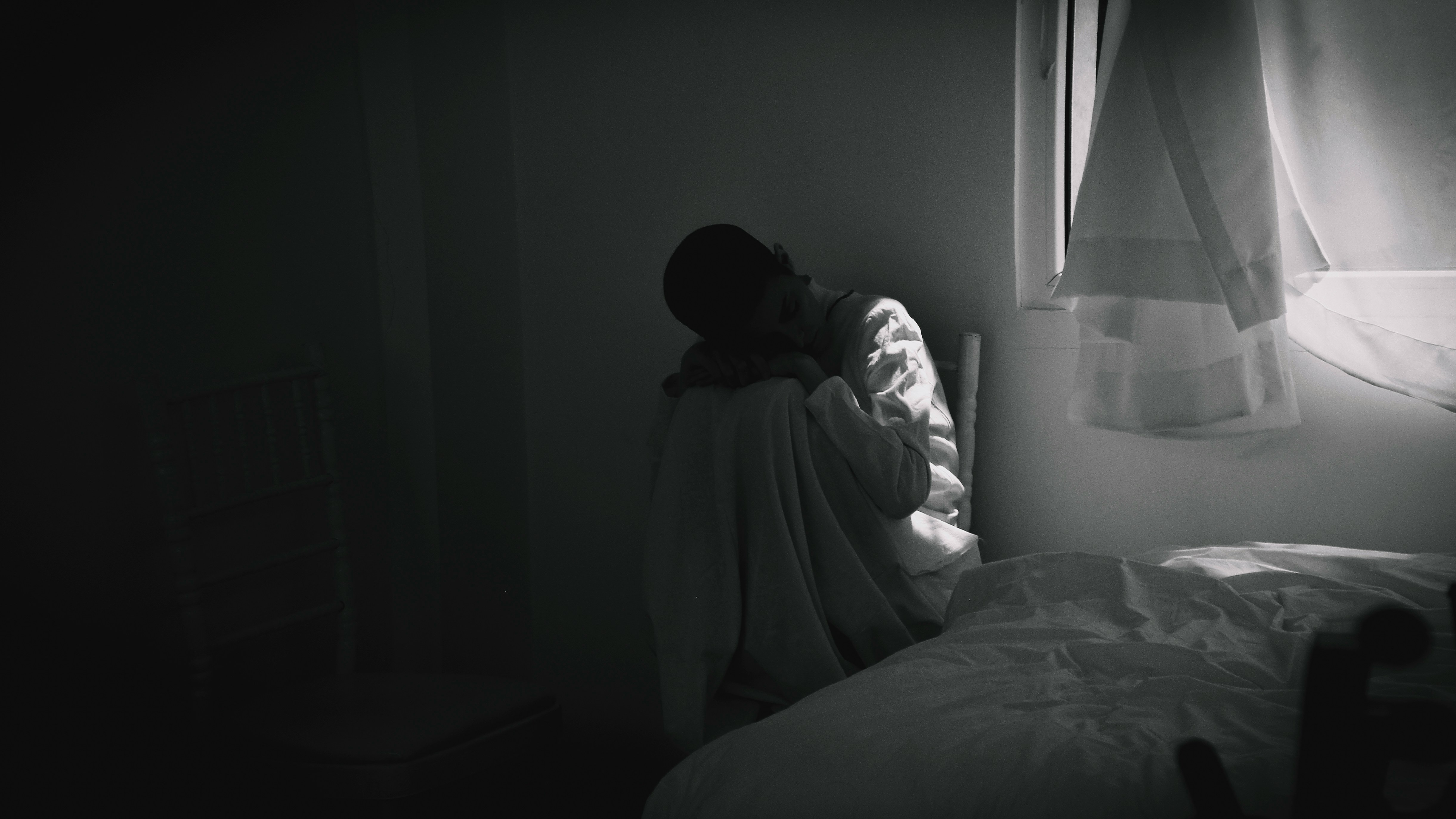Einsame Person sitzt zusammengesunken im dunklen Zimmer neben einem Fenster – symbolisiert emotionale Belastung und Einsamkeit im Studium.