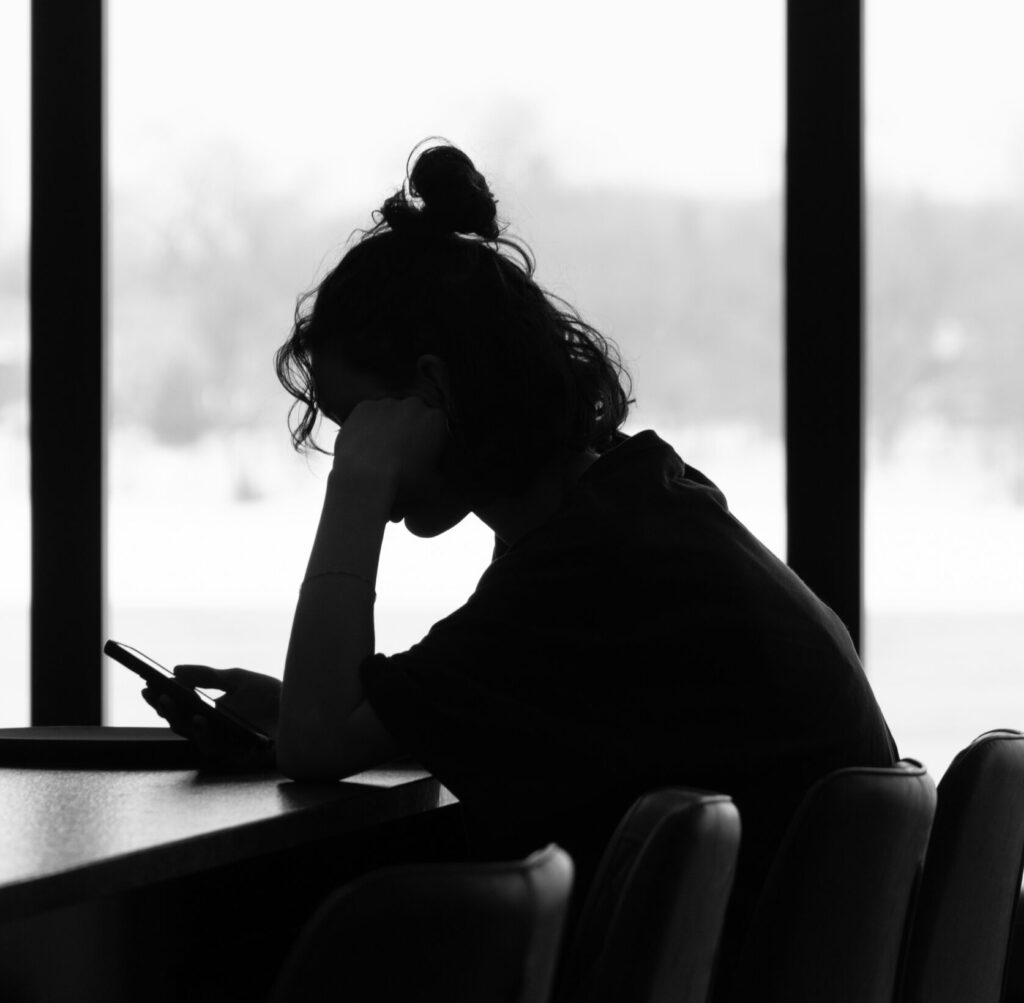 Junge Person sitzt allein an einem Tisch, blickt auf ihr Smartphone, den Kopf in die Hand gestützt; hinter ihr helle Fenster mit verschwommenem Blick ins Freie. Symbolbild für Stress.